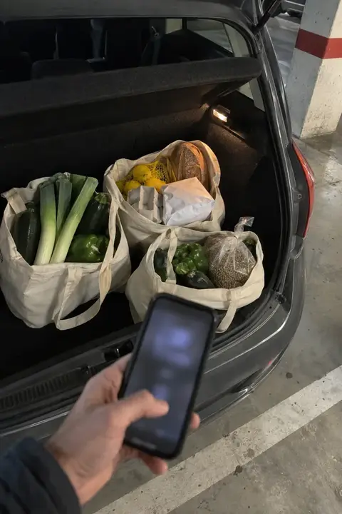 Maletero de un coche con bolsas de tela llenas de verdura, pescado y lentejas, y una mano con el móvil